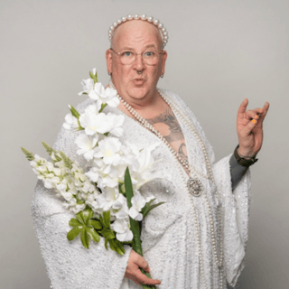 Eine Person trägt ein glitzerndes weißes Kleid und hält einen Blumenstrauß. Sie trägt eine Perlenkette und und Perlenkrone.
